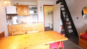 Una cocina compacta con armarios de madera, microondas y un lavamanos pequeño. Hay una mesa de comedor con sillas, y una escalera que lleva a una zona superior con estanterías.