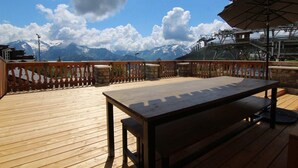 Terraza de madera con mesa y bancos, con vistas a un paisaje montañoso que incluye un telesilla.