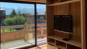 A spacious balcony with a wooden railing, a television on a wooden stand, and a view of mountains and buildings.