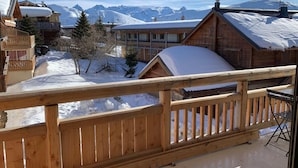 A wooden balcony with a view of snow-covered mountains and a chalet-style building.