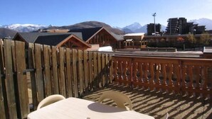 Una terraza de madera con una mesa y sillas, con vistas a un paisaje montañoso y edificios al fondo.