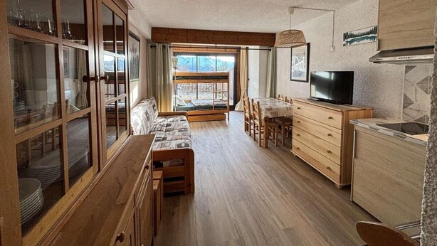 Amplia sala de estar con un banco de madera, una mesa de comedor y una zona de cocina con televisor y alacenas.
