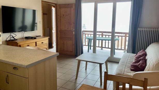 A cozy room with a wooden ceiling, a TV on a wooden cabinet, a small table, a sofa, and a balcony with a view. - 30m from ski slopes, 1st floor, balcony, tv, ski locker, 32m², Alpe d'Huez (L'Alpe d'Huez)