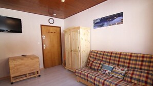A room with a wooden cabinet, a plaid sofa, a television mounted on the wall, and a door with a clock. - 5th floor, tv, 16m², Alpe d'Huez (L'Alpe d'Huez)