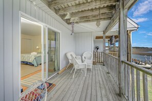 Eine Holzterrasse mit weißen Korbstühlen, eine Glastür, die zu einem Schlafzimmer führt, und Blick aufs Wasser.