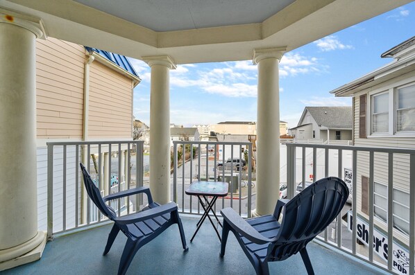 A balcony with two chairs and a small table, overlooking a street with buildings and parked cars. - Belmont Towers 307 by Seaside Vacations & Sales (Ocean City)
