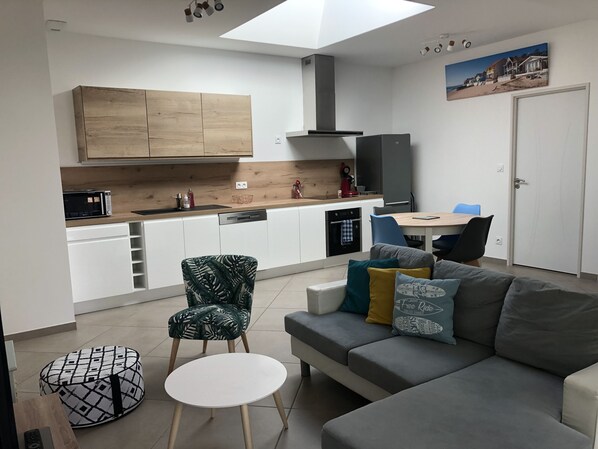 A modern kitchen with white cabinets, a wooden countertop, and a built-in oven. There is a dining area with a table and chairs, and a gray sofa with colorful cushions.