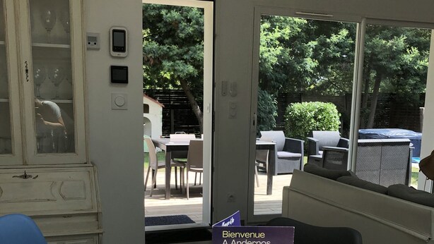 A reception area with a wooden table, a framed welcome sign, a cabinet with glass doors, and a view of the outdoor patio with furniture. - 800 M from the basin / direct access to cycle path 6 people (Andernos-les-Bains)