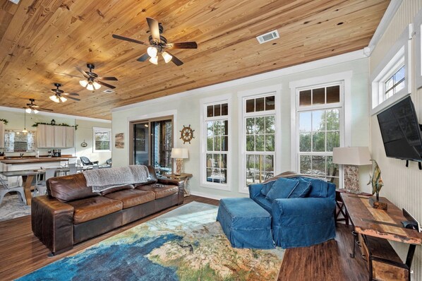 A spacious living room with a wooden ceiling, large windows, a leather sofa, a blue armchair, and a dining area with a wooden table.