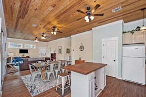 A spacious living area with a wooden ceiling, a dining table with chairs, a kitchen area with a refrigerator, and a small island with a sink.