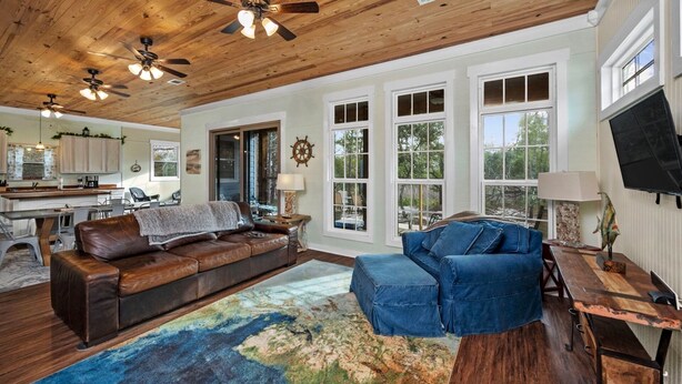 Un salón espacioso con techo de madera, ventanas amplias, un sofá de cuero, un sillón azul y un comedor con mesa de madera.