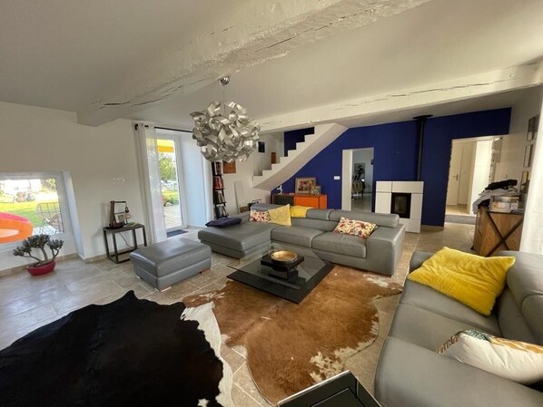 A spacious living room with a large gray sofa, a coffee table, and a cowhide rug. There is a staircase leading to another level, and a bookshelf filled with books.