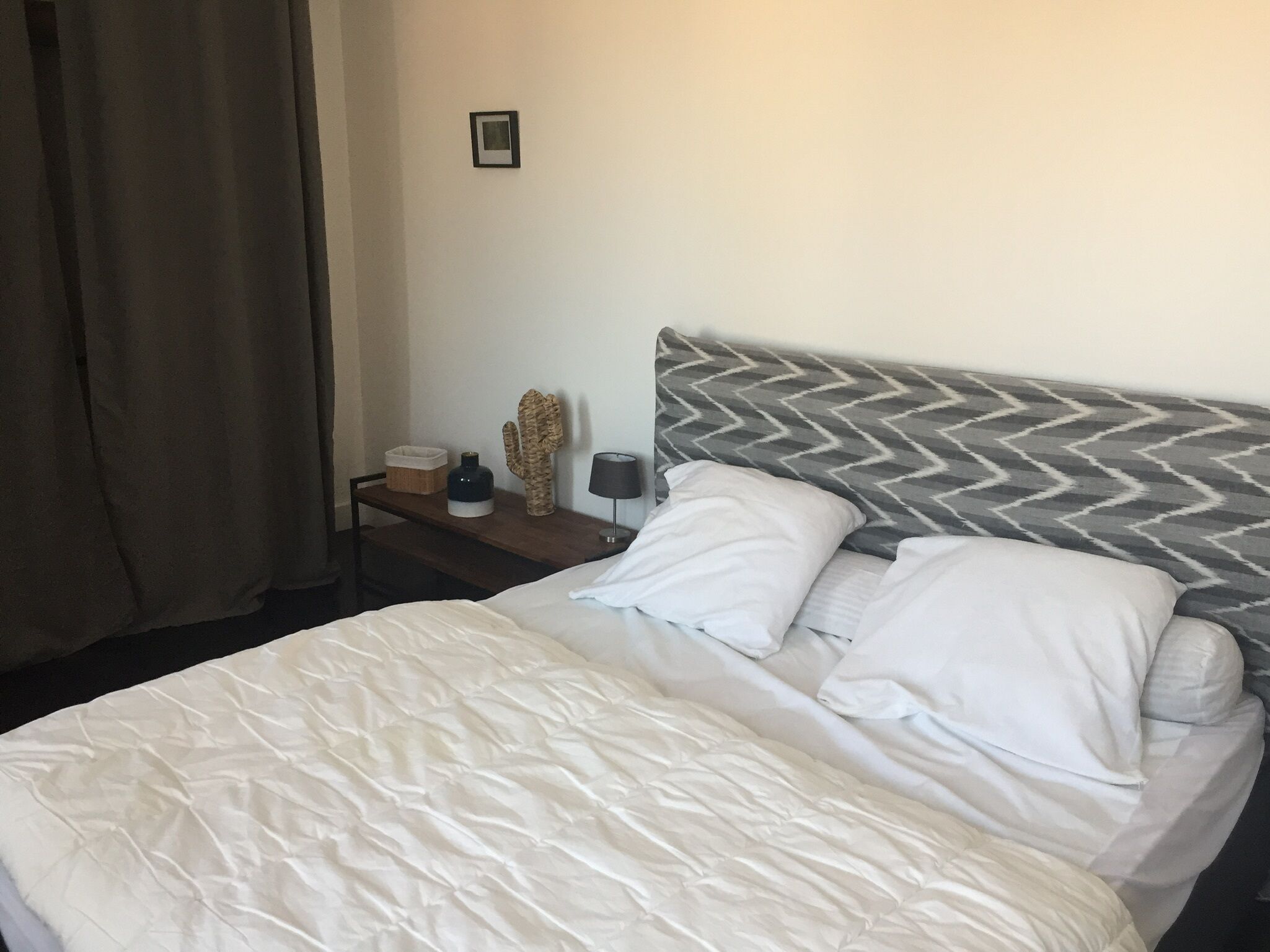 Un lit bien fait, avec du linge de lit blanc, une table de chevet en bois surmontée d’une lampe, un cactus décoratif et un tableau encadré accroché au mur.