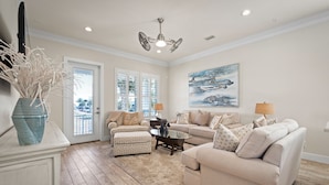 Un salon spacieux avec un canapé beige, un pouf à motifs et une table basse. Un tableau orne le mur, un ventilateur de plafond est installé au plafond et une porte donne accès à un balcon.