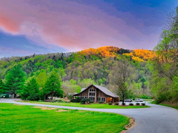 A rustic cabin with a large porch, surrounded by lush green fields and a forested hill in the background. - Golden Eagle Landing: Modern Mountain Getaway, Hot Tub, Pet Friendly, WiFi, Long (Ferguson)