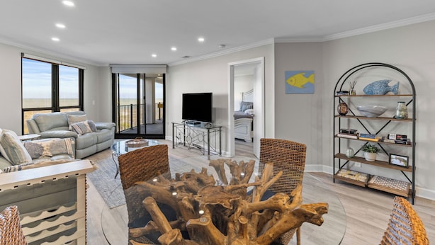 A modern living room with a large window, a flat-screen TV, and a wooden coffee table. - Summer House 204 - Pool/Beach - Ocean View! (Isle of Palms)