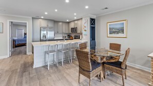 A modern kitchen with white cabinets, a central island, and a dining area with wicker chairs and a glass table. - Summer House 204 - Pool/Beach - Ocean View! (Isle of Palms)