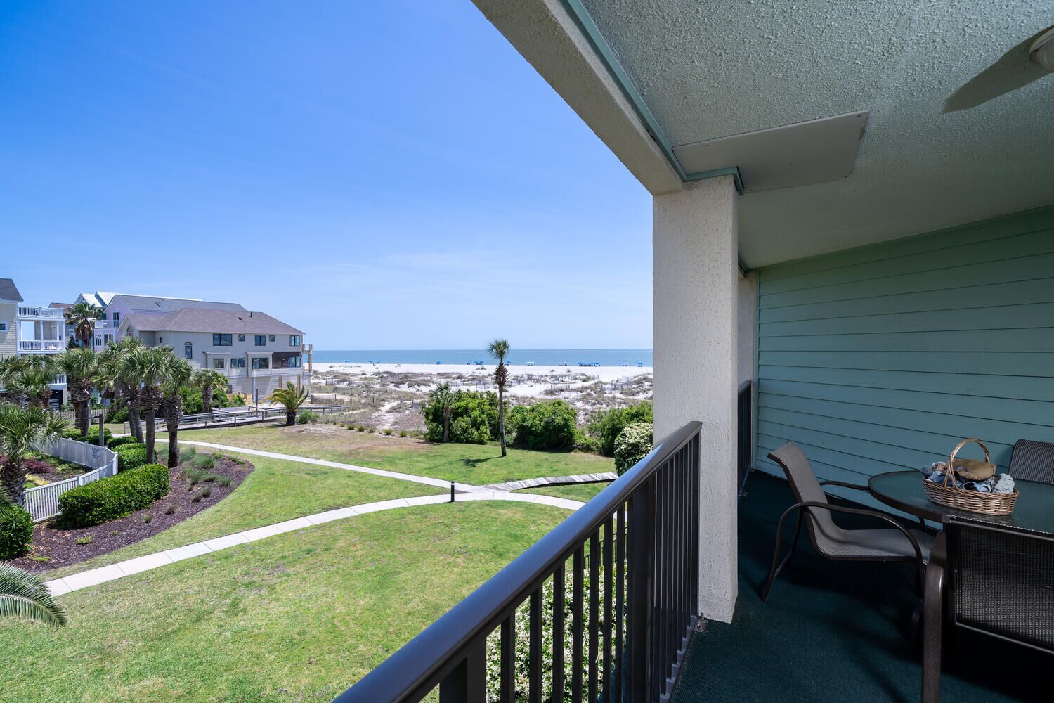 A balcony with a view of the beach, a grassy area, and a building with a balcony.