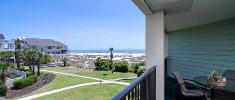 A balcony with a view of the beach, a grassy area, and a building with a balcony.