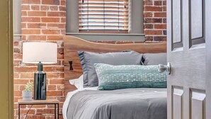 A bedroom with a brick wall, a wooden headboard, a bedside table with a lamp, and a window with blinds.