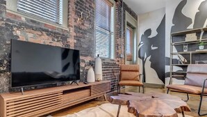 Un salon moderne avec un grand téléviseur à écran plat posé sur un meuble en bois, des fauteuils en cuir, une table basse ronde et un mur en briques agrémenté de grandes fenêtres.