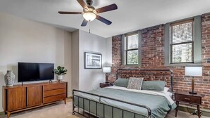 Une chambre avec un mur en briques, un lit avec un cadre en métal, un téléviseur posé sur un meuble en bois et un ventilateur de plafond.