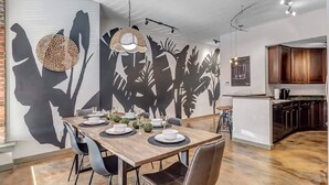 Un espace de salle à manger moderne, avec une table en bois dressée pour quatre personnes, entourée de chaises noires. Un grand mur est décoré d’une fresque représentant un feuillage tropical et des silhouettes.