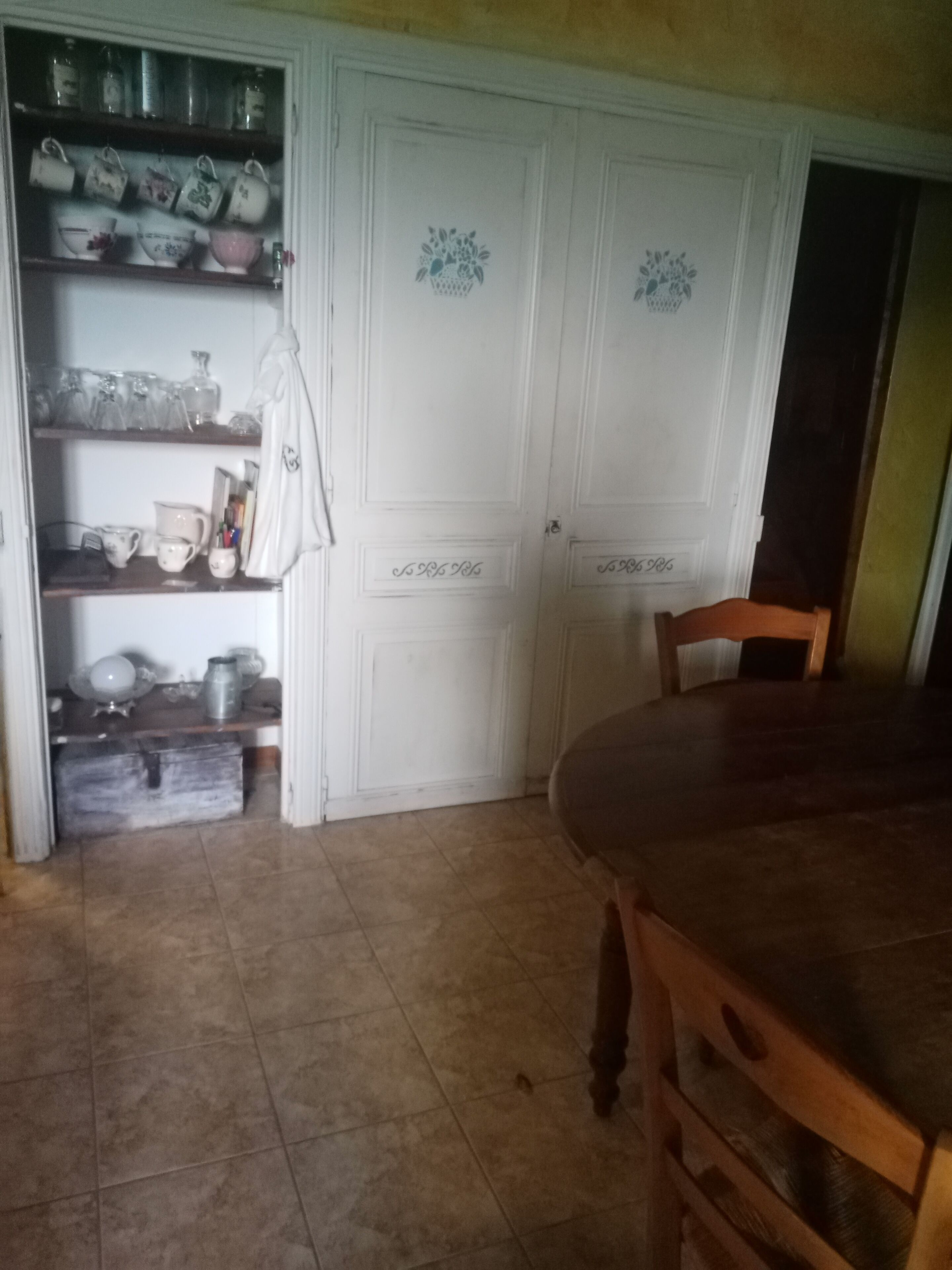 Una cocina con una mesa y sillas de madera, un armario con utensilios de cocina y una puerta blanca con motivos florales decorativos.