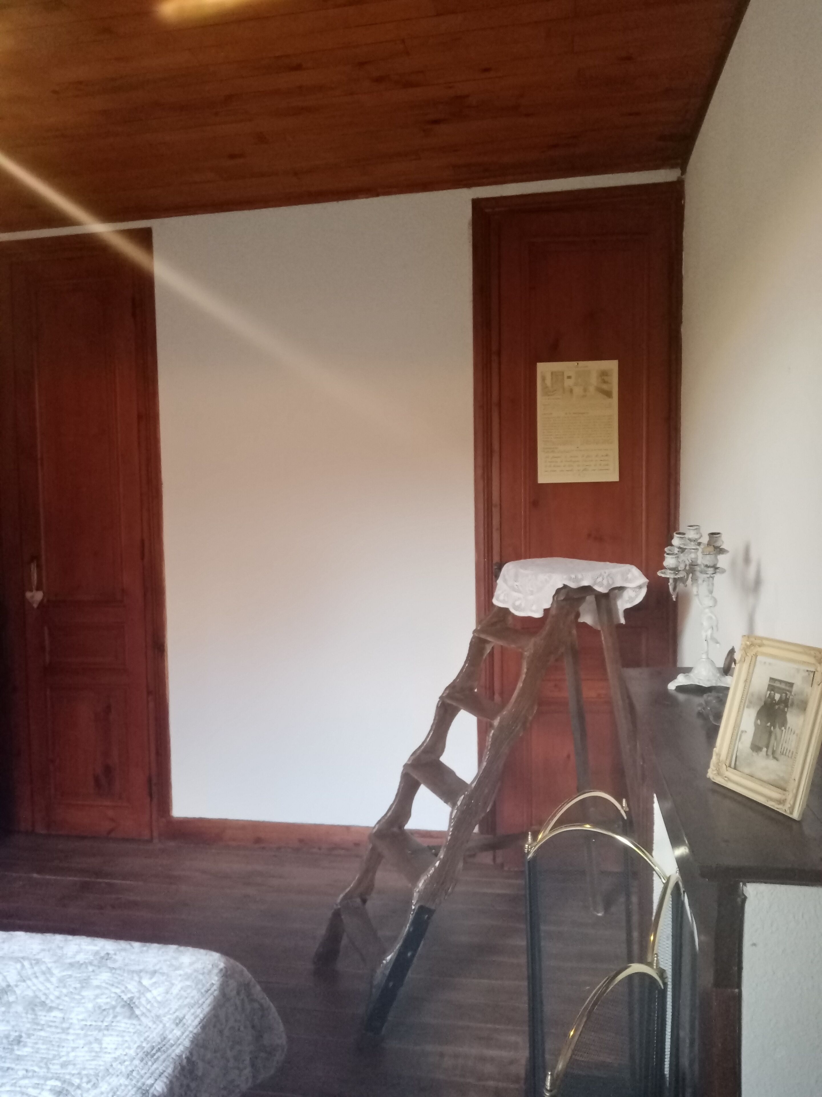A room with a wooden ladder leaning against a wall, a white table with a lace tablecloth, and a candelabra on a wooden dresser.