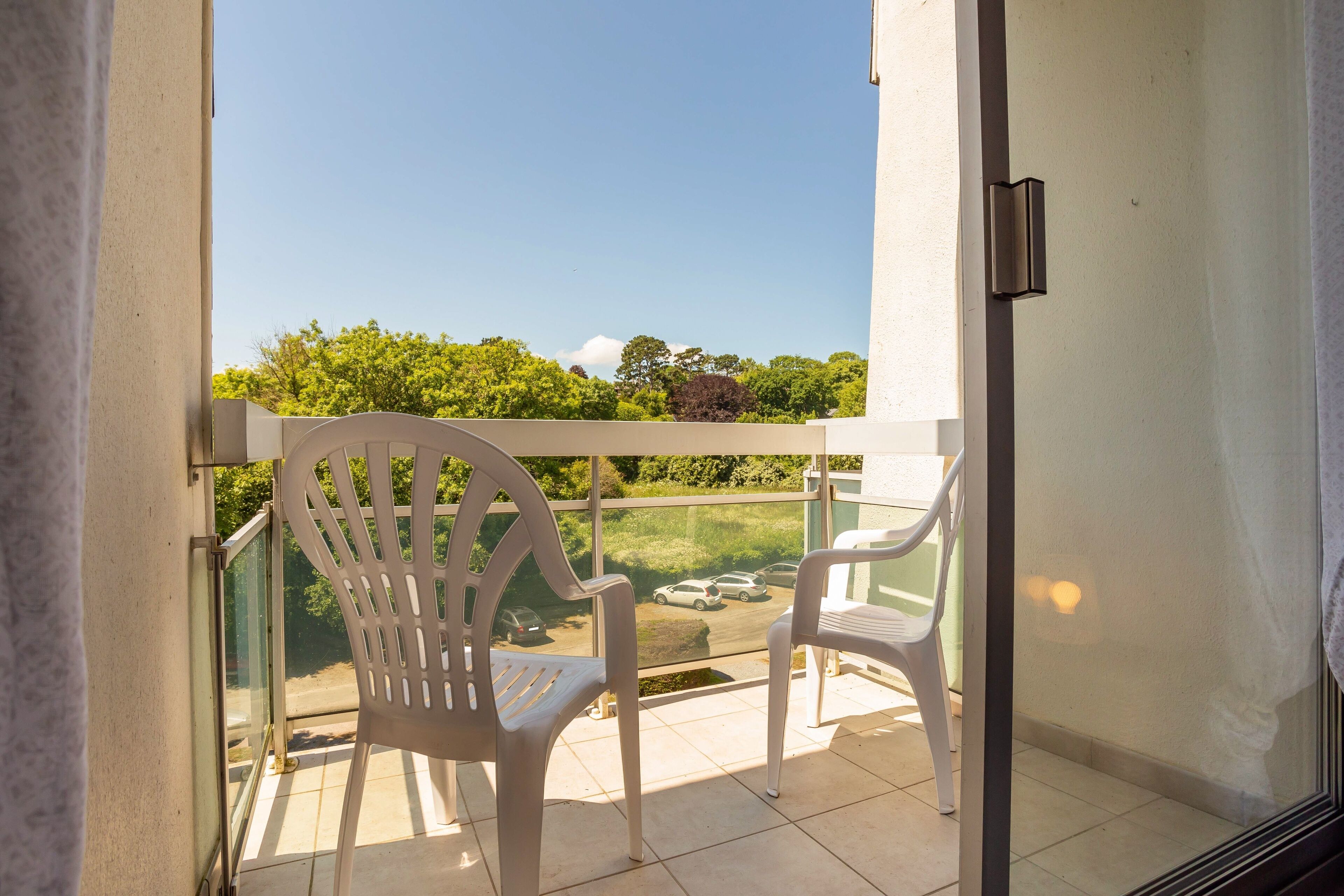 A balcony with two white plastic chairs, a glass railing, and a view of a parking area and greenery.