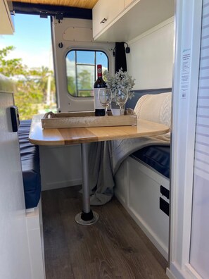 Un intérieur de camping-car compact comprenant un coin cuisine, un espace repas avec une table en bois et une fenêtre donnant sur un paysage verdoyant.