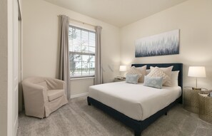 Ein Schlafzimmer mit einem Bett, zwei Nachttischen, einem Sessel, einem Fenster mit Vorhängen und einem an der Wand hängenden Gemälde.