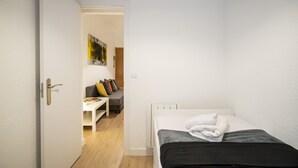 Una habitación pequeña y ordenada con una cama, una puerta blanca y vistas a otra habitación con un sofá y una mesa.