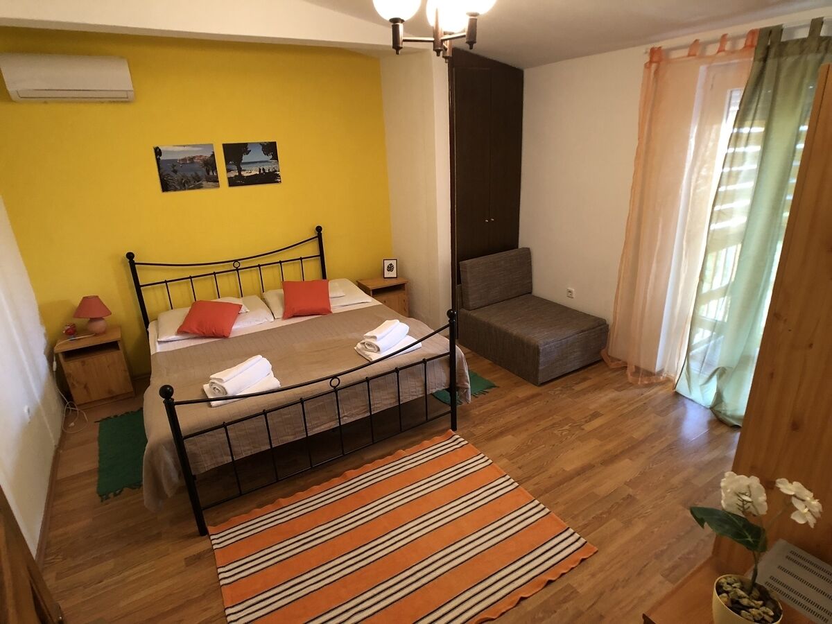A bedroom with a bed, a bedside table, a lamp, a sofa, a window with curtains, and a wall-mounted air conditioner.