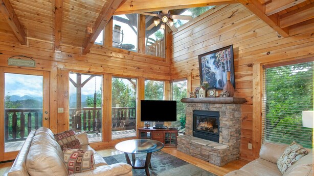 A cozy cabin interior with a stone fireplace, leather sofas, a flat-screen TV, and large windows offering scenic views. - Bella Yani - Hot Tub, Pool Table, & Air Hockey! (Sevierville)
