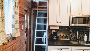 Una cocina compacta con armarios blancos, un microondas y una escalera que lleva a una zona de descanso en un altillo.