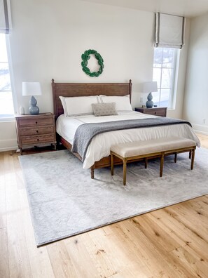 A bedroom with a wooden bed, a bench, two bedside tables with lamps, a nightstand, a green wreath on the wall, and a large window.