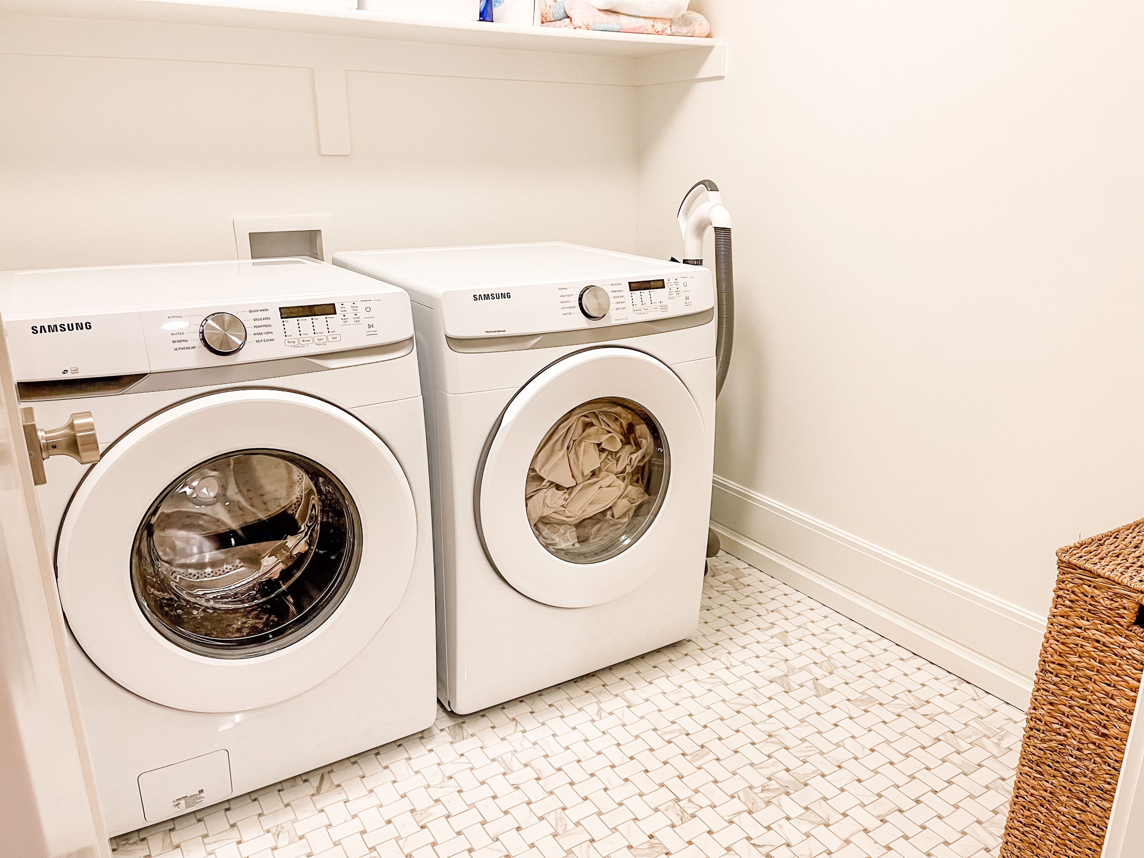 Deux lave-linge à chargement frontal Samsung blancs, dans une salle de bain carrelée avec un mur blanc.
