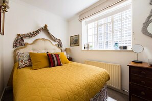 A neatly arranged bedroom with a large bed, a window with blinds, a dresser, and a bedside table with a lamp.