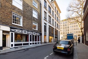 A street view with a black car parked on the side, a building with a modern facade, and a tree with bare branches.