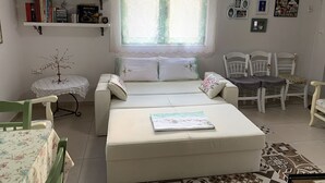 A cozy room with a bed, a small table, and a chair. There is a window with curtains, a bookshelf, and a wall-mounted air conditioner.