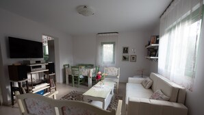A living room with a TV, entertainment unit, floral sofa, dining table, chairs, and a window with curtains.