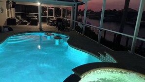 A swimming pool with a hot tub, surrounded by a covered patio area with seating and a view of the water. - SW CAPE CORAL: 5 BR 4 BA CANAL HOME with BOAT DOCK  w/heated / lit pool, jacuzzi (Cape Coral)