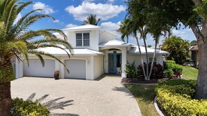 A two-story house with a white exterior, a blue front door, and a driveway leading to a garage. - SW CAPE CORAL: 5 BR 4 BA CANAL HOME with BOAT DOCK  w/heated / lit pool, jacuzzi (Cape Coral)
