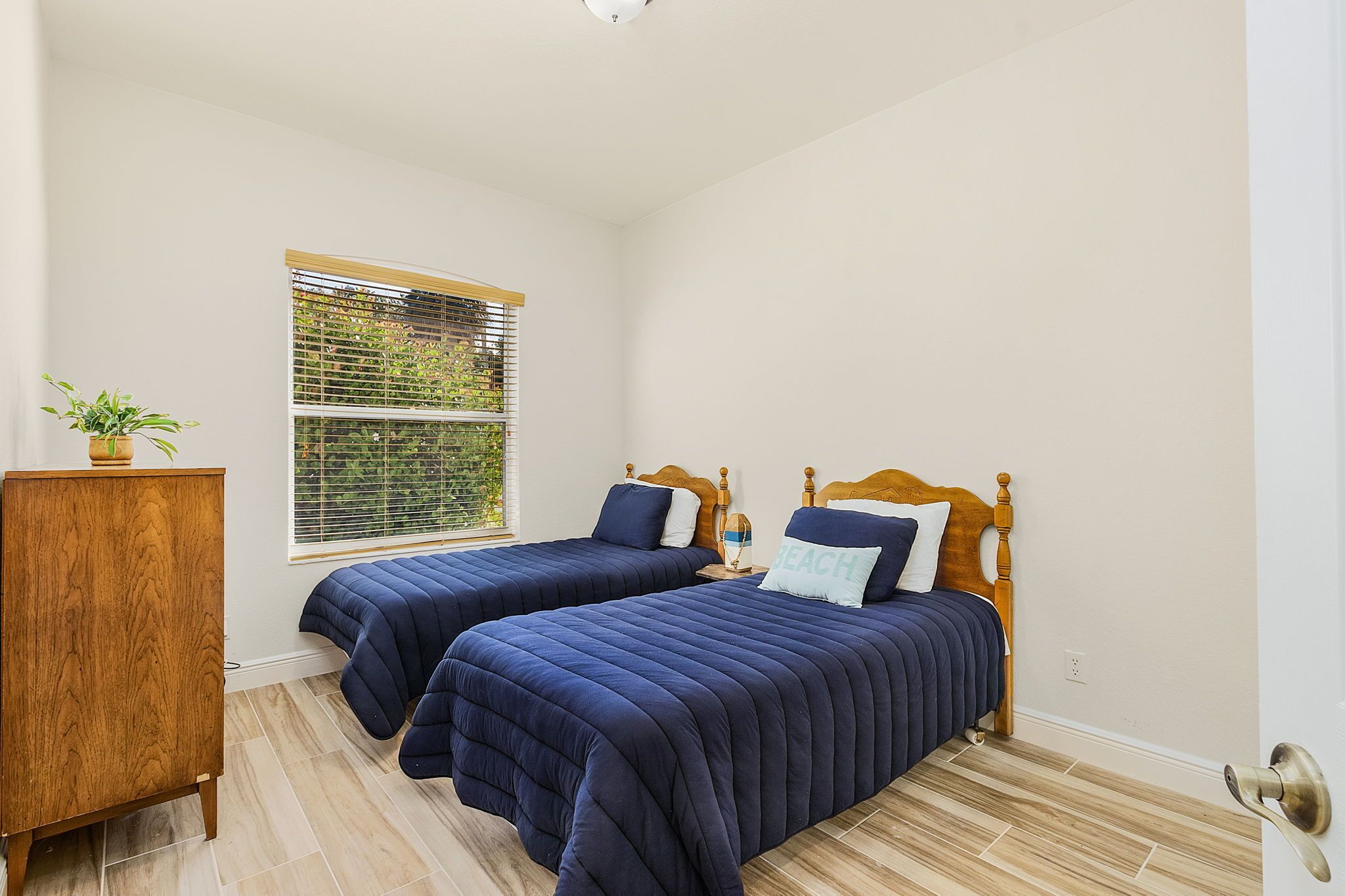 A bedroom with two beds, a wooden dresser, and a window with blinds.