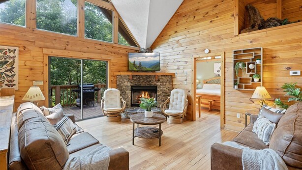 A cozy cabin interior with a fireplace, wooden walls, and a large window overlooking a forest.