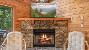 A stone fireplace with a fire, flanked by wicker chairs, in a wooden cabin with a TV above the fireplace.