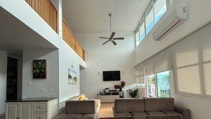 A spacious living area with a high ceiling, a ceiling fan, a flat-screen TV mounted on the wall, a kitchen area with a countertop, and a large window providing natural light.
