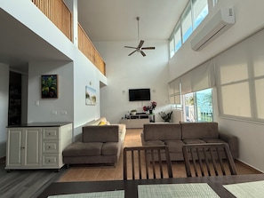 A spacious living area with a high ceiling, a ceiling fan, a flat-screen TV mounted on the wall, a kitchen area with a countertop, and a large window providing natural light.