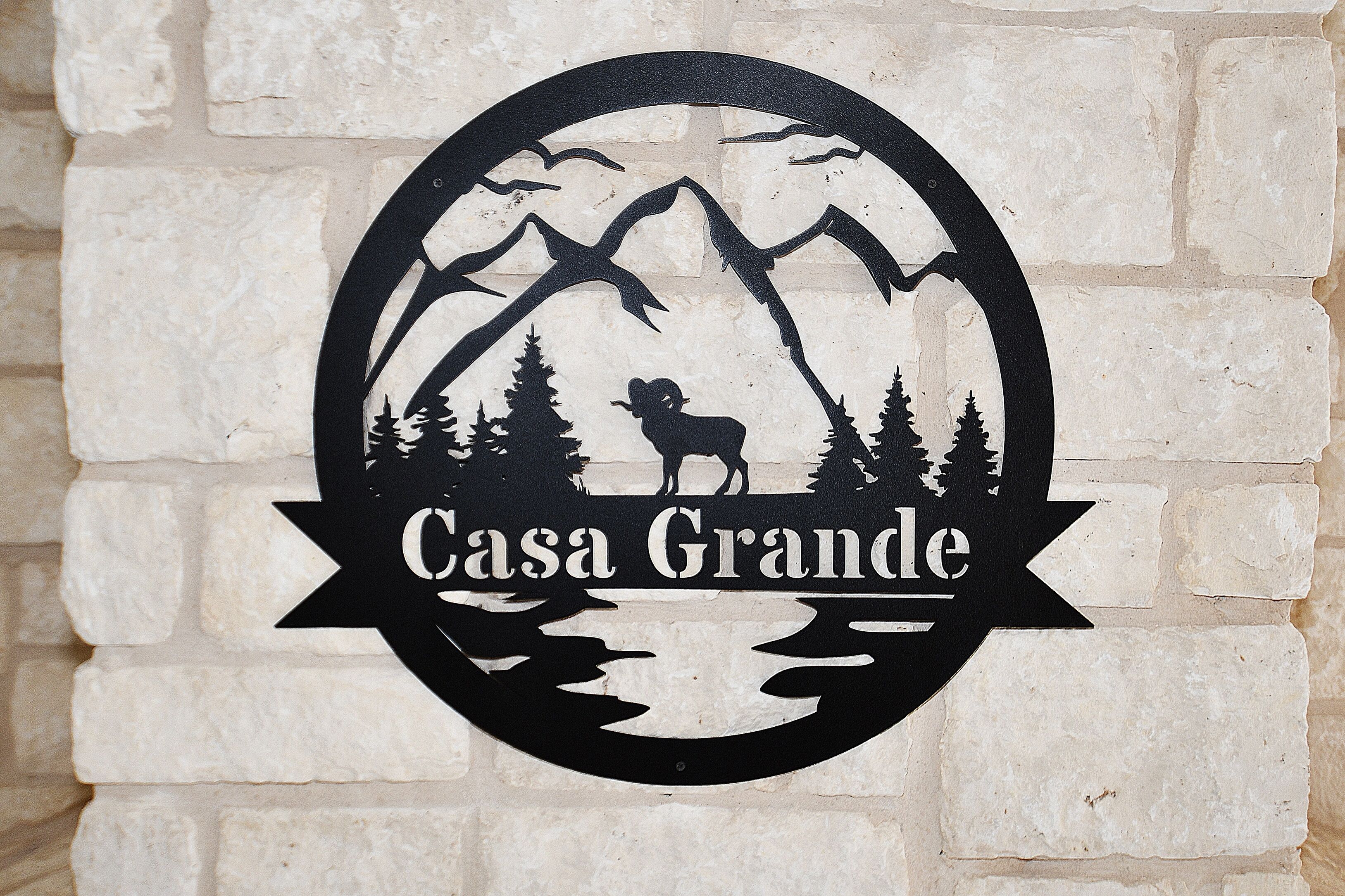 Uma placa preta com silhuetas de montanha e árvore, e o texto 'Casa Grande' em uma parede de tijolos brancos.